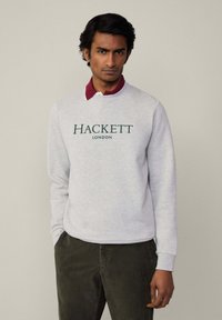 Šedá mikina s logom "HACKETT LONDON" v zelenej farbe, golier v burgundskej farbe, dlhé rukávy, jemná textúra. Nosí sa s koordinovanými tmavými nohavicami.