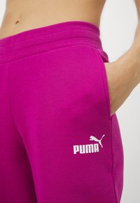 Fuksjowe spodnie sportowe wykonane z miękkiego materiału, z elastycznym pasem, z bocznymi kieszeniami i białym logo Pumy na dolnej części nogawki.