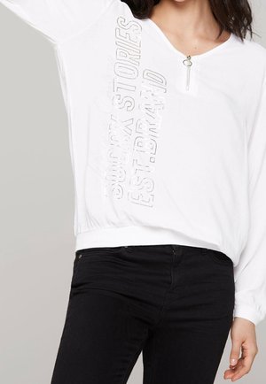 Kobieta w białej koszuli z długim rękawem z suwakiem przy dekolcie i pionowym napisem "SOUL ROCK STORIES EST. BRAND" w połączeniu z czarnymi spodniami.