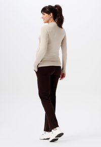 Beige Langarmshirt im Slim-Fit, kombiniert mit dunkelbraunen Hosen. Die Hose verfügt über Seitentaschen und einen leichten Schlag am Saum. Weiße Sneakers.
