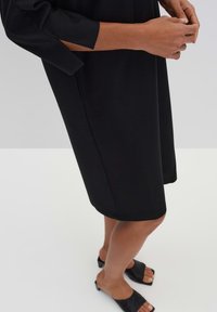Schwarzes knielanges Kleid aus glattem Stoff. Mit Dreiviertelärmeln und lockerer Passform, kombiniert mit schwarzen offenen Slip-On Sandalen.