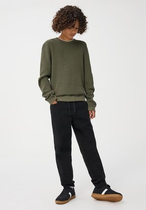 Zielony sweter dzianinowy o prążkowanej strukturze, z okrągłym dekoltem, w połączeniu z czarnymi spodniami dresowymi z troczkami. Czernie buty sportowe z gumową podeszwą.