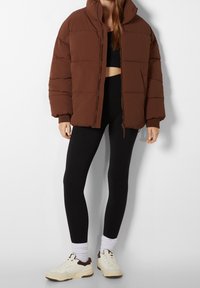 Giacca puffer marrone con colletto alto, caratterizzata da sezioni trapuntate. Abbinata a leggings neri e sneakers crema con dettagli marroni.