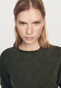 Mørkegrøn V-hals sweater lavet af en tekstureret strik, båret over en mørkere undertrøje. Model med glat hår og neutralt udtryk.