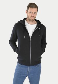 Haut à capuche zippé noir avec cordon de serrage, poches avant, poignets et ourlet côtelés, porté sur un t-shirt blanc, présenté sur un fond blanc.