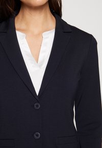 Navy blazer with notched lapels, two buttons, and side pockets, layered over a white satin blouse with a V-neck design. Smooth texture.