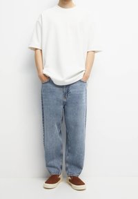Persona vestida con una camiseta blanca de manga corta y holgada, jeans de corte recto de color azul claro y zapatillas de tela marrones, de pie con las manos en los bolsillos.