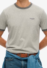 Man in een lichtgrijze SuperDry Athletics T-shirt met een donkergrijze kraag en mouwranden, een bruine leren riem, blauwe jeans en een vastgebonden blauw overhemd.