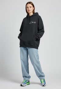 Felpa nera con cappuccio e tasca frontale, con scritte bianche in corsivo. Abbinata a jeans azzurri e sneakers sportive multicolori.