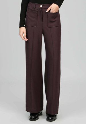 Femme portant un pantalon marron foncé à taille haute et jambes larges avec poches plaquées à l'avant, une fermeture à bouton et zip, associé à des chaussures et un haut noirs.