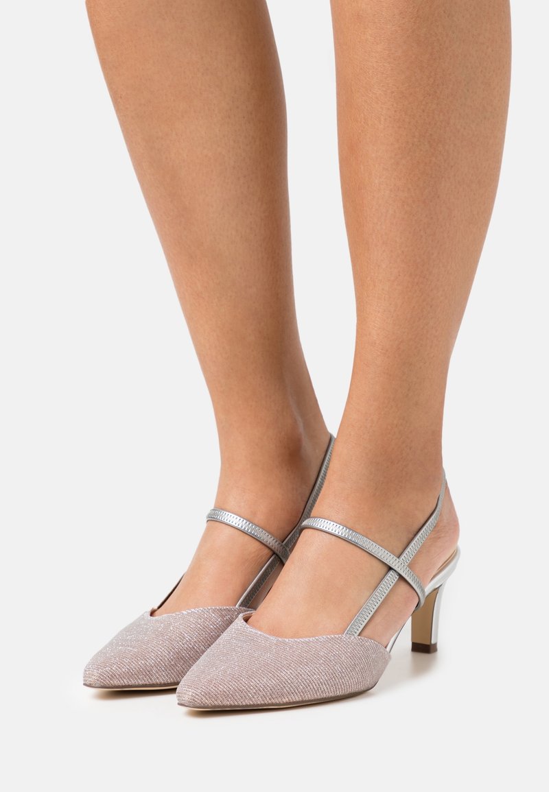 Femme portant des escarpins slingbacks rose pâle brillant à bout pointu, avec des brides argentées et des talons blocs de hauteur moyenne sur un fond clair.