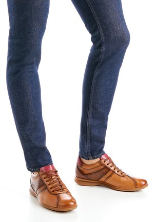 Braune Ledersneaker mit roten Akzenten, einem flachen Profil, abgerundeter Spitze und kontrastierenden Streifen an den Seiten, kombiniert mit dunklen Jeans.