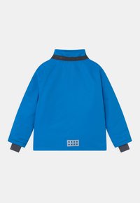 Blå vattentät jacka med hög krage, svarta krage accenter och elastiska ärmslut. Har en silverfärgad rektangulär logotyp på baksidan.