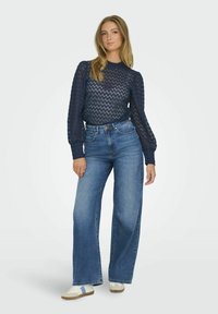 Pull en tricot à motif bleu marine, à manches longues, associé à un jean en denim taille haute et à jambes larges. Chaussures : baskets blanches avec des accents bleus.