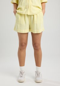 Completo corto giallo chiaro con righe blu verticali. Il tessuto appare testurizzato, con una camicia larga con bottoni e pantaloni corti.