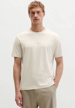 Young man with blond hair wearing a light beige short-sleeve t-shirt and beige pants, standing against a plain white background.