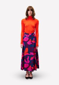Femme debout portant un pull roulé orange vif et une longue jupe colorée avec des motifs floraux abstraits roses, rouges et bleus, chaussures noires.