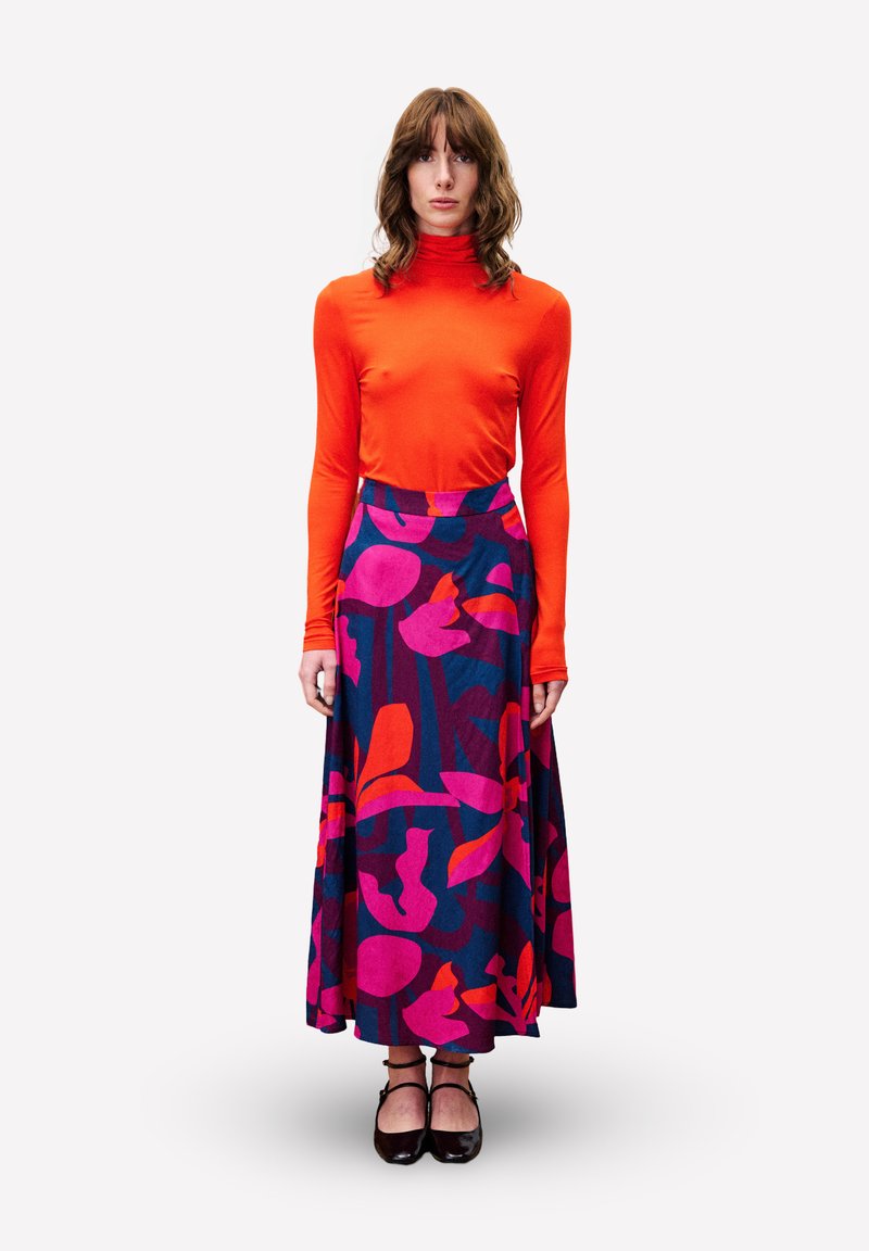 Femme debout portant un pull roulé orange vif et une longue jupe colorée avec des motifs floraux abstraits roses, rouges et bleus, chaussures noires.