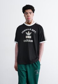 Melna īsām piedurknēm adidas T-kreklā ar baltām akcentiem, kurā ir uzraksts "Originals Dept." un zīmols. Savienots ar zaļām sporta biksēm.