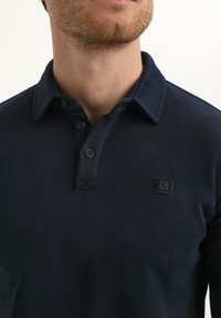 Navyblauwe polo met een gestructureerde stof, een drieknoopsluiting en een klein zwart logo op de linkerbovenkant.