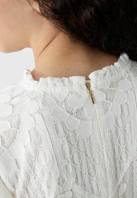 Witte kant blouse met een textuurpatroon, ruches bij de halslijn en een gouden ritsluiting aan de achterkant. Glad materiaal met ingewikkelde details.