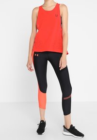 Red tank top with mesh detailing, paired with black leggings featuring orange accents and striped patterns. Black athletic shoes complete the look.