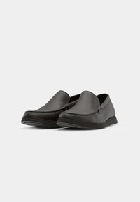 Paire de mocassins en cuir gris foncé, type slip-on, avec bouts arrondis et semelles noires sur un fond clair uni.