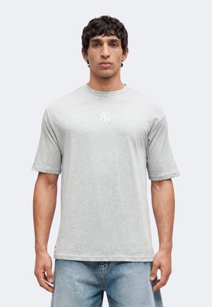 Mand iført lysegrå T-shirt med korte ærmer og lille hvidt New York Yankees-logo, kombineret med lyseblå cowboybukser mod ensfarvet baggrund.