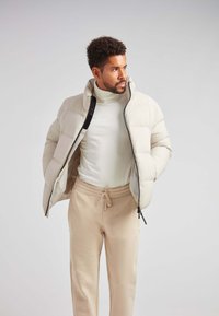 Beige pufferjakke med høj krave og lynlås, båret over en cremefarvet turtleneck og lyse beige sweatpants. Blød tekstur, minimalistisk design.
