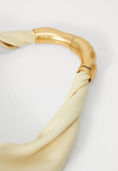 Κρέμ τσάντα δερμάτινη με περιστροφικό λουράκι και χρυσές λεπτομέρειες, διαθέτοντας καμπυλωτό χερούλι και λεπτομέρεια λογότυπου κοντά στο σημείο πρόσδεσης.