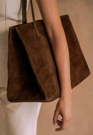 Person wearing white pants carries large brown suede tote bag over arm against beige background.
