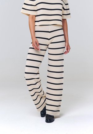 Henkilö, jolla on beigenväriset neulotut housut ja niiden kanssa sopiva lyhythihainen yläosa, jossa mustat vaakaraitoja, yhdistetty mustiin kenkiin, seisoo valkoisella lattialla.