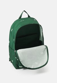 Grön ryggsäck med dragkedjestängning. Har en slät, texturerad tyg, invändig ficka och Nike-logotyper tryckta på utsidan.