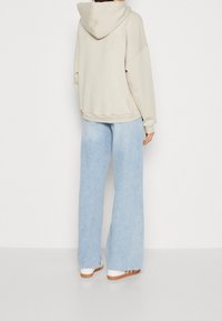 Béžová oversized mikina s kapucí, v kombinaci se světle modrými širokými džínami. Materiály vypadají měkce a mají uvolněný střih.