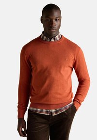 Oranžový pletený sveter s rebrovaným lemom a manžetami, ktorý je vrstvený cez károvanú košelu. Sveter má na hrudi malé vyšívané logo.