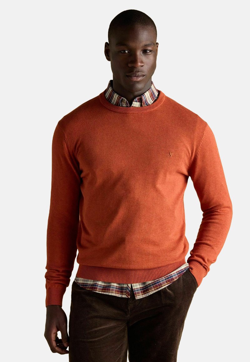 Oranžový pletený sveter s rebrovaným lemom a manžetami, ktorý je vrstvený cez károvanú košelu. Sveter má na hrudi malé vyšívané logo.