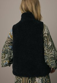 Donker, zacht, mouwloos fleecevest met een hoge kraag, gedragen over een kleurrijk, patroonrijk shirt met lange mouwen en golvende ontwerpen.