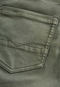 Olive rohelise värviga kangas tasku kontrastsete õmblustega, millel on eritellimusel chevron muster klapi peal. Sile tekstuur ja puhtad servad.