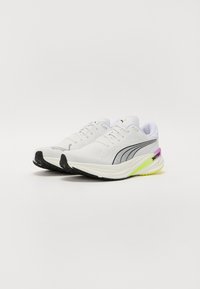 Zapatillas deportivas blancas con material de malla y sintético, con acentos en negro y púrpura, suela texturizada y un detalle en el talón amarillo.