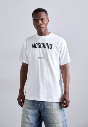 Valge puuvillane T-särk, millel on silmapaistev must Moschino logo ja allpool "Milano" tekst. Särgil on laxd ja lühikesed varrukad.