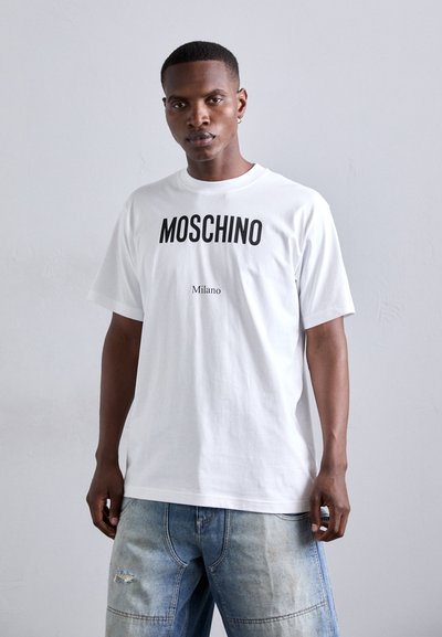 Bijela pamučna majica s istaknutim crnim Moschino logom i tekstom "Milano" ispod. Majica ima opušteni kroj i kratke rukave.