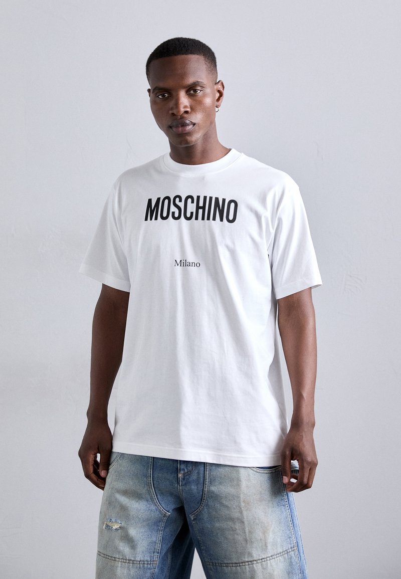 Balts kokvilnas T-krekls ar izteiktu melnu Moschino logo un tekstu "Milano" zem tā. Krekls ir brīvā griezumā un ar īsām piedurknēm.