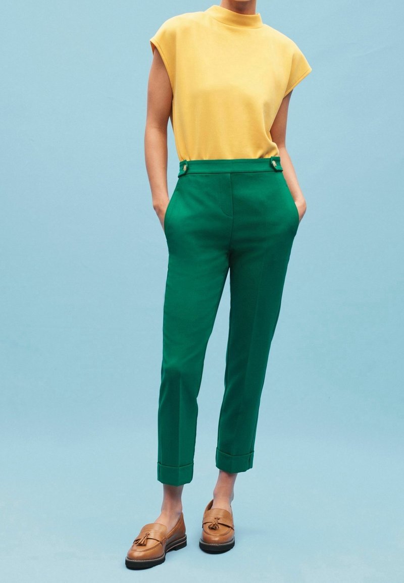 Personne portant un haut sans manches jaune, un pantalon vert taille haute avec des ourlets retournés, et des mocassins marron à glands sur un fond bleu clair.