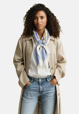 Femme aux cheveux bouclés portant un trench-coat beige, une chemise blanche, une écharpe bleue à motifs nouée autour du cou, et un jean bleu, les mains dans les poches.