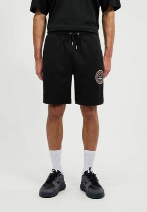 Schwarze Baumwollshorts mit elastischem Bund und verstellbarem Kordelzug. Mit einem Logopatch am Oberschenkel. Kombiniert mit schwarzen Sneakern und weißen Socken.