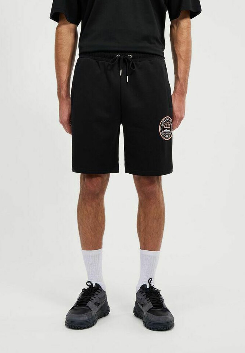 Pantalones cortos de algodón negros con cintura elástica y cordón ajustable. Presentan un parche con el logo en el muslo. Combinados con zapatillas negras y calcetines blancos.