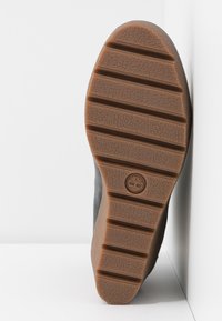Semelle en caoutchouc marron avec des rainures horizontales et un logo d'arbre au centre, faisant partie d'une chaussure noire appuyée contre une surface blanche.