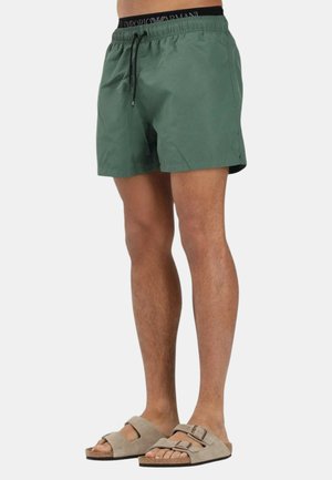 Uomo che indossa pantaloncini da bagno verdi con coulisse nera e fascia con logo, abbinati a sandali beige a doppio cinturino, in piedi davanti a uno sfondo bianco.