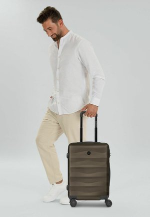 Uomo con camicia bianca e pantaloni beige che trascina una valigia rigida nera con quattro ruote e manico esteso.