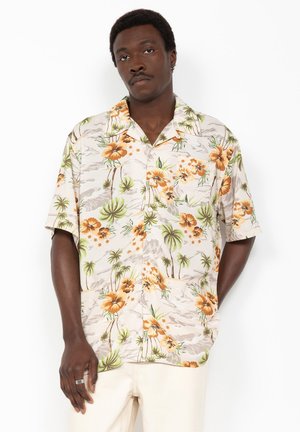 Homme portant une chemise beige à manches courtes avec des boutons, ornée d'imprimés d'hibiscus orange et de palmiers verts, associée à un pantalon de couleur claire.